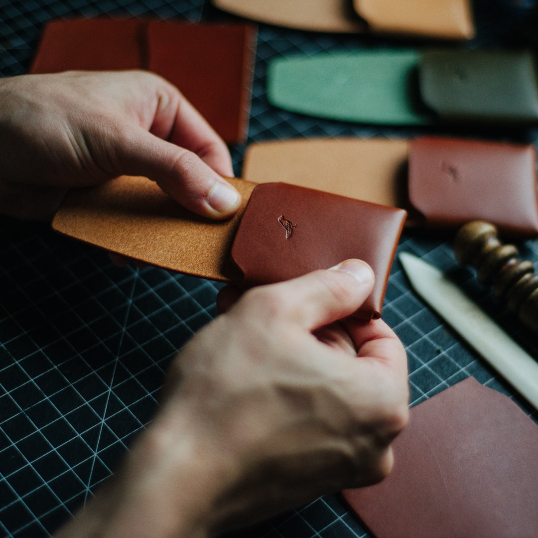 Crafting a Brown Cash Emerson Wallet: A man is seen working on the wallet against a backdrop featuring other color variations of the product and various crafting tools