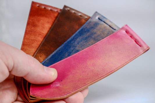 Four bookmarks in colors: red, blue, dark brown, light brown, held in the hand of a man with a gray background