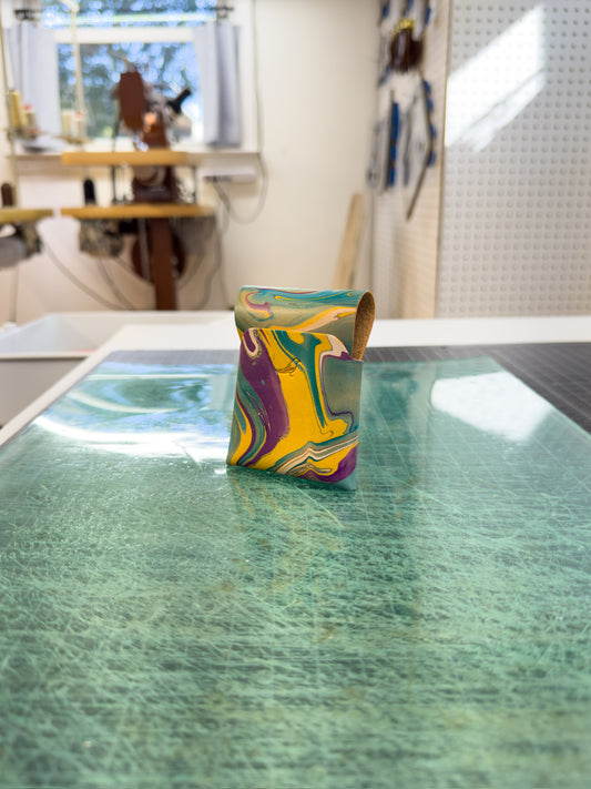 Mardi Gras 1 leather wallet placed on a green surface, viewed from the front and back, with a workspace visible in the background where similar wallets are being created