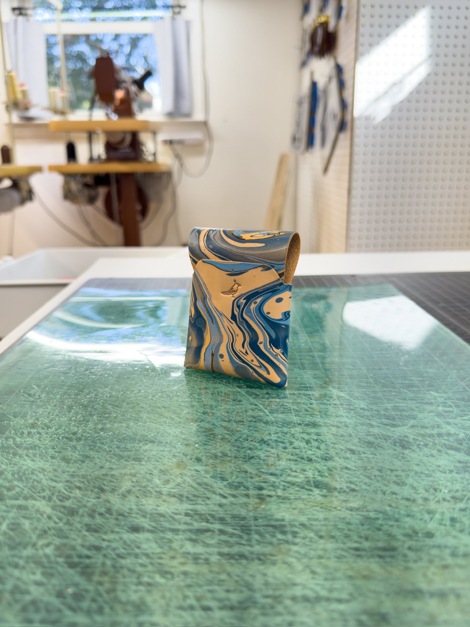 Blue leather wallet placed on a green surface, viewed from the front and back, with a workspace visible in the background where similar wallets are being created