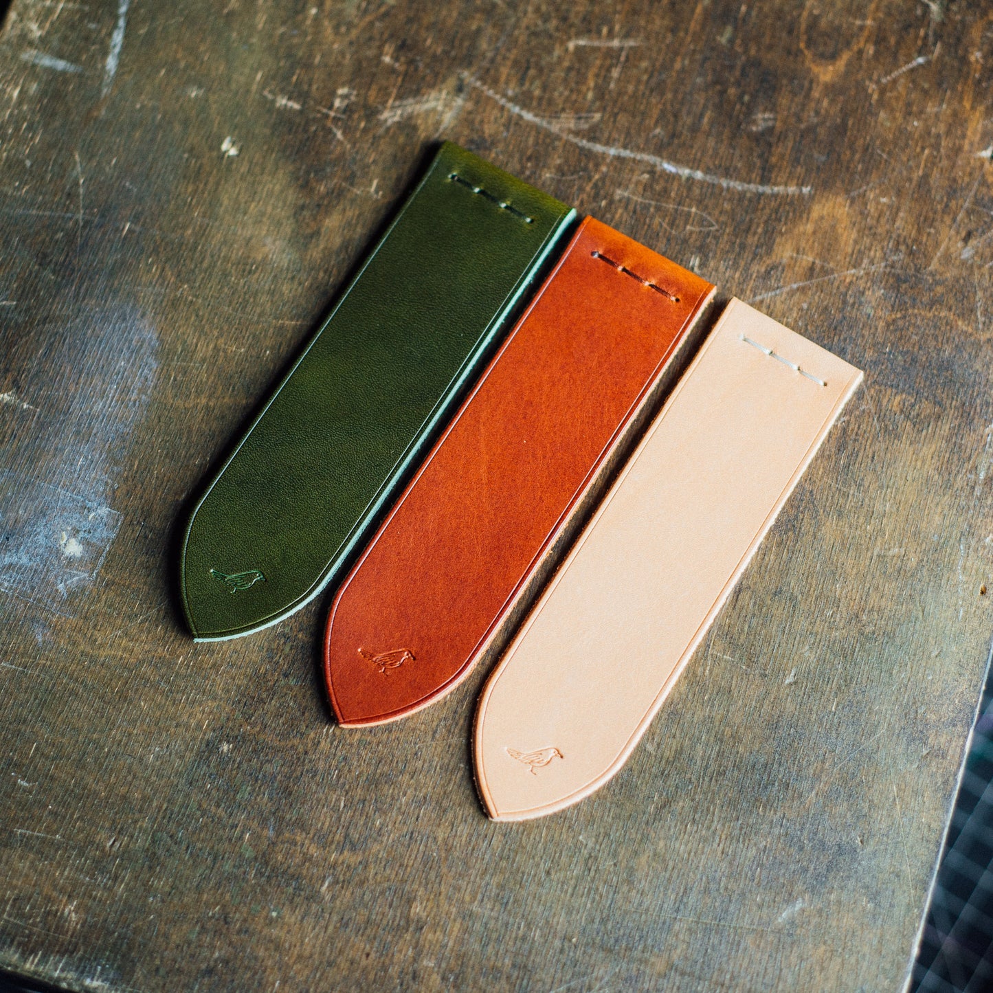 Elegant full-grain leather bookmarks meticulously hand-stitched from premium Buttero leather. On a green table, showcasing the craftsmanship of olive, chestnut, and natural-colored bookmarks