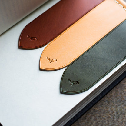 Elegant hand-stitched full-grain leather bookmarks made from premium Buttero leather. Resting on the pages of a book atop a brown table, showcasing olive, chestnut, and natural-colored bookmarks