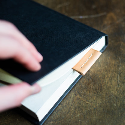 Exquisite full-grain leather bookmark meticulously hand-stitched from luxurious Buttero leather. Placed into an open book by a man, positioned on a crafting table