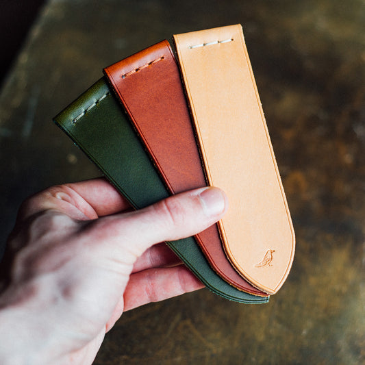 Elegant full-grain leather bookmarks meticulously hand-stitched from premium Buttero leather. Held by a hand against a background of a green table, showcasing the craftsmanship of olive, chestnut, and natural-colored bookmarks