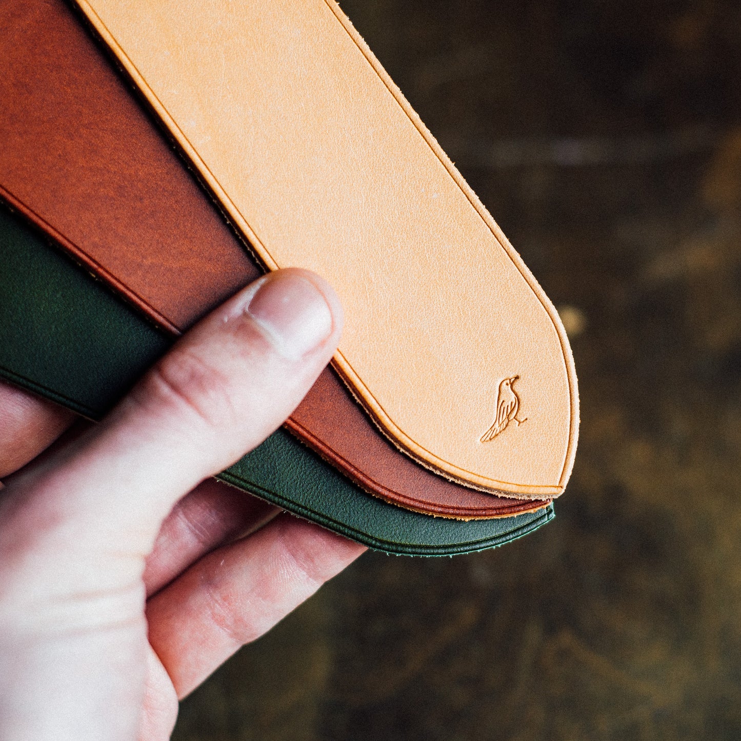 Elegant full-grain leather bookmarks meticulously hand-stitched from premium Buttero leather. Held by a hand against a background of a green table, showcasing the craftsmanship of olive, chestnut, and natural-colored bookmarks